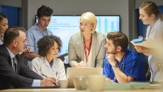 People standing and sitting around a table with a screen showing a graph behind them People standing and sitting around a table with a screen showing a graph behind them