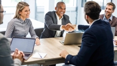 Five businesspersons sitting around a table with two of them shaking hands Five businesspersons sitting around a table with two of them shaking hands