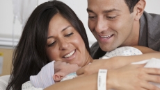 One person holding a baby while lying down with another person looking over their shoulder at the baby