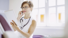Healthcare provider sitting in an office wearing a stethoscope and looking at a tablet Healthcare provider sitting in an office wearing a stethoscope and looking at a tablet