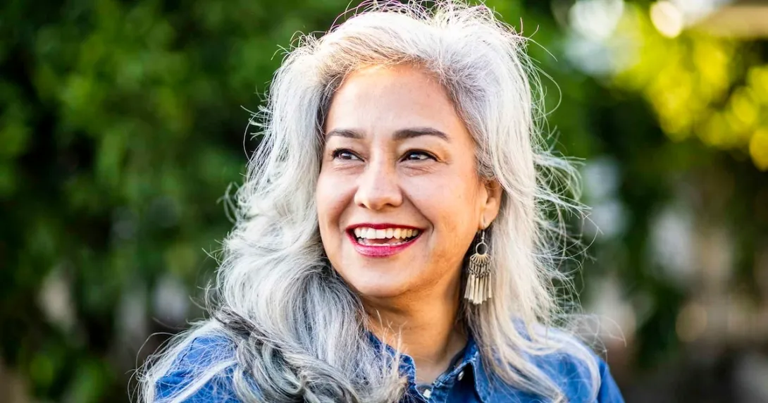 Person with long gray hair smiling while standing in front of trees