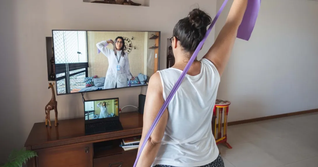 A woman watching and copying exercises with a resistance band in her living room, guided by a physical therapist online