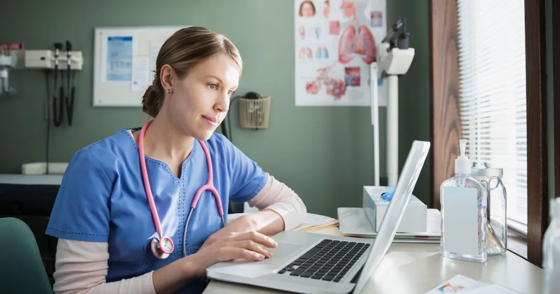 Healthcare provider wearing a stethoscope around their neck and scrubs sitting at a desk and looking at a computer