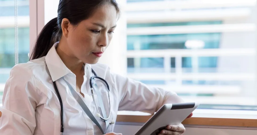 Healthcare provider sitting near a window while wearing a stethoscope and looking at a tablet