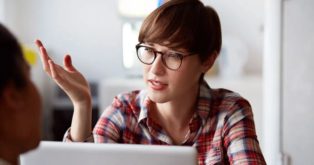Person wearing glasses and a plaid shirt sitting at a computer with their hand up as though talking to someone on the computer