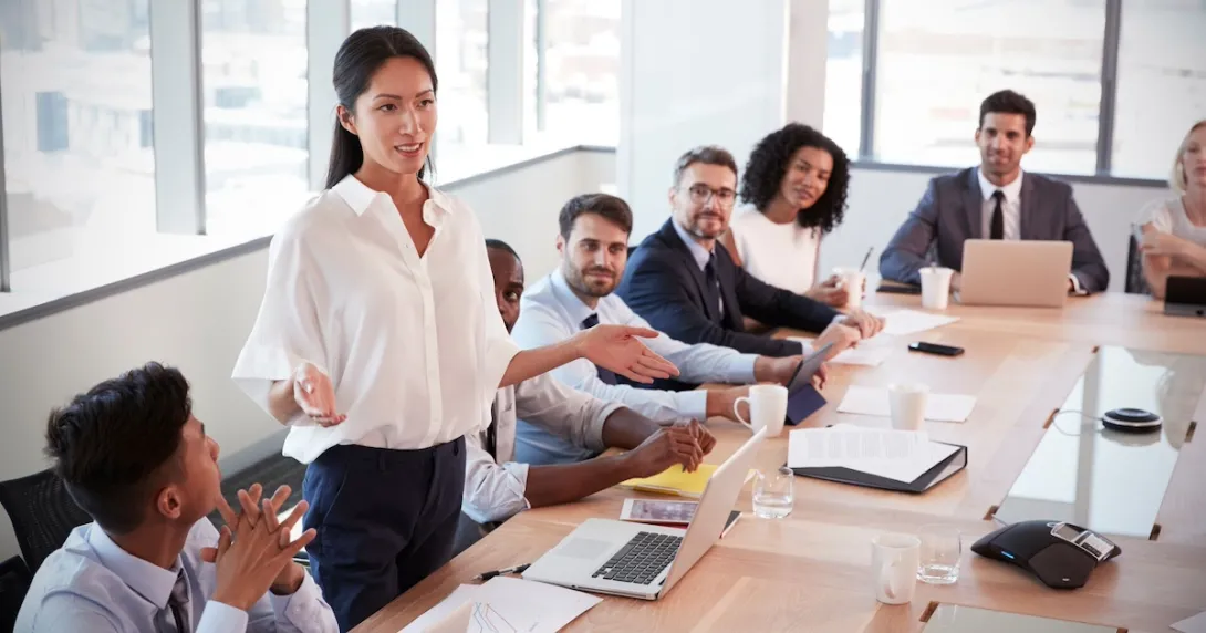 Numerous people in a board room sitting around a table with one person standing up